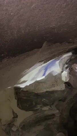 A close-up, low-angle shot pans through a dimly lit cave-like environment. The rough, textured ceiling of the cave is visible, appearing dark brown and granular. Below the ceiling, a section of lighter-colored rock or material is illuminated, creating a bright, stark contrast. A thin, blue stripe is visible on this illuminated surface, appearing to be painted or stained. The rest of the cave interior consists of dark, shadowed rock formations with irregular shapes and textures. The lighting is focused on the illuminated section, casting deep shadows throughout the rest of the scene. The overall impression is one of a subterranean space with geological features and a single, deliberate mark.