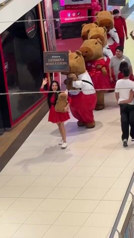 A procession walks through a brightly lit commercial space with white tiled floors. Leading the group is a woman in a red dress, holding a teddy bear, and waving. Behind her is a group of four costumed characters resembling capybaras. Some are wearing red and white outfits. A sign held by the group reads "LLEVAMOS A MAMA A EUROPA" (We are taking Mom to Europe). There are several men in the procession walking alongside and behind the characters. A man in a gray polo shirt pulls a rolling suitcase. Various shops and signs are visible in the background.