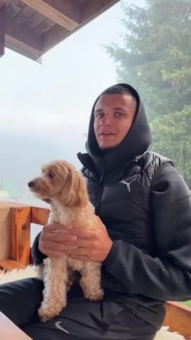 A young man with a black hooded jacket and black pants is holding a small, light brown, curly-haired dog on his lap. The man is smiling and looking towards the camera. The dog is looking to the left of the frame. They are sitting outdoors on a wooden bench with a sheepskin rug on it, likely on a porch or balcony. Behind them, a misty forest with trees is visible. The weather appears to be foggy or overcast.