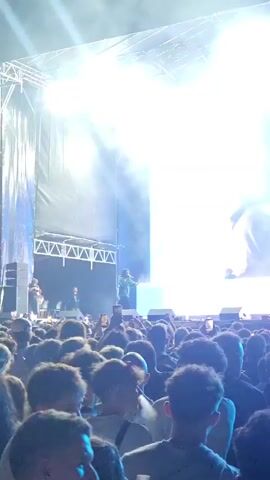 A live concert is taking place. A performer is on stage under bright blue lights, facing a large crowd. A massive screen behind the performer displays a close-up of the performer's face. The crowd is packed together, with many people holding up their phones to record the event. The atmosphere appears energetic and engaging.