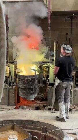 In an industrial setting, a foundry worker, equipped with protective gear including a face shield, apron, and leg coverings, stands near a large, smoking crucible. The crucible is positioned in front of a tilted furnace, with a reddish glow emanating from within. The crucible appears to contain molten material, generating significant heat and white smoke. The worker observes the process. The surroundings include industrial equipment and brick walls.