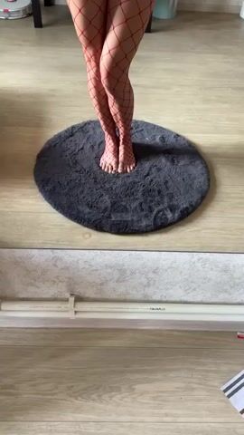 A woman stands barefoot on a round grey rug in a room. She is wearing red fishnet stockings and red underwear. She is holding a smartphone in her right hand and taking a selfie. She has a tattoo on her left thigh. In the background, there is a dresser with various items on it, a vacuum cleaner, and a window. The walls are light-colored. The woman adjusts her underwear. She puts on a pair of black cat ears. She poses for the camera.