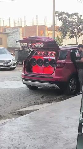 The video displays a street scene with a customized red SUV parked on the side of a paved road. Its rear hatch is open, revealing an elaborate car audio system installed in the trunk. The system features multiple large subwoofers (at least four visible), several mid-range speakers, and tweeters, all integrated into a custom red enclosure. Additional circular lights or speakers are mounted on the interior of the open tailgate.

An adult male, wearing a dark t-shirt and pants, stands next to the red SUV, looking down at a device that appears to be a laptop or tablet in his hands. He seems to be operating or adjusting the audio system, from which loud music is playing.

In the background, a silver sedan with the license plate "D3H-692" is parked. Another red car briefly drives past further down the street. The setting appears to be a daytime street in an urban or suburban area, with buildings, a power pole, and some foliage visible under a bright sky.