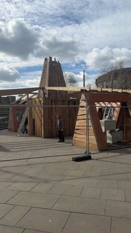 The video shows a construction site with a wooden structure under construction. The structure appears to be an abstract building with multiple angular sections and a central tower-like element made of vertical wooden planks. A metal fence surrounds the construction area. Two workers wearing safety helmets can be seen near the structure, one of them is carrying a ladder. Another person wearing a brown jacket walks past the construction site. In the background, a historic church building can be seen. The ground is covered with gray paving stones. The sky is partly cloudy.