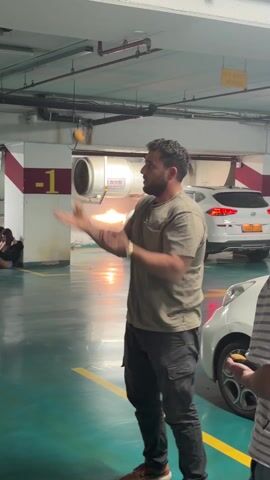A man is juggling three small, brown objects in an indoor parking garage. The man is wearing a short-sleeved olive-green t-shirt and dark pants, and has a wrist watch. He is standing on a green parking space in between two parked white cars. In the background, on the left, two people are sitting on the floor. Behind the man is a ventilation system attached to the ceiling, which has a red and white painted post next to it. The parking garage is otherwise mostly white.