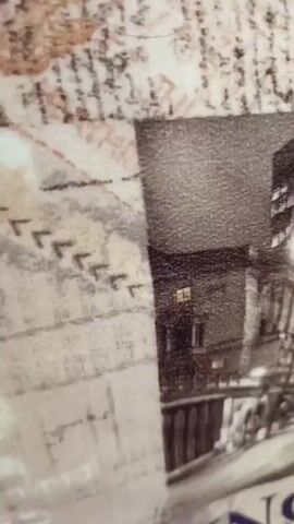 The video shows a close-up, angled view of a collage or print that depicts a nighttime cityscape. In the foreground, a red double-decker bus is prominently displayed, facing the viewer. Behind the bus, a large, modern building with many illuminated windows is visible. To the left and right of the bus, there are glimpses of older, classical architecture with columns. The overall color scheme is grayscale with the exception of the red bus. The background appears to be a textured surface with what looks like text or script elements. The lighting is dim, suggesting an evening or nighttime scene. No people are clearly discernible, though there might be faint figures in the distance in the architectural areas.