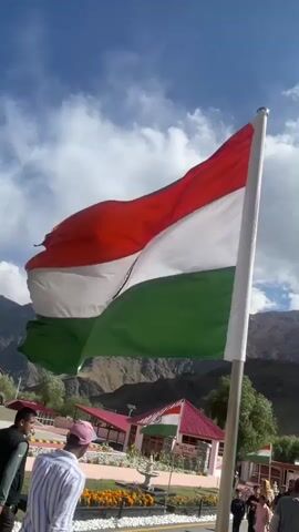 A vertical shot captures the Indian flag waving in the wind against a backdrop of a blue sky with scattered clouds and distant mountains. The flag, with its saffron, white, and green stripes and the Ashoka Chakra in the center, dominates the foreground. In the middle ground, several people are visible, some standing and observing, while others are walking. Behind them, a building with a red roof and an Indian flag displayed prominently is visible. The overall environment appears to be an outdoor public space, possibly a tourist spot or a memorial, with manicured flower beds in the foreground. The video is a short, static shot with no discernable action or significant interaction.