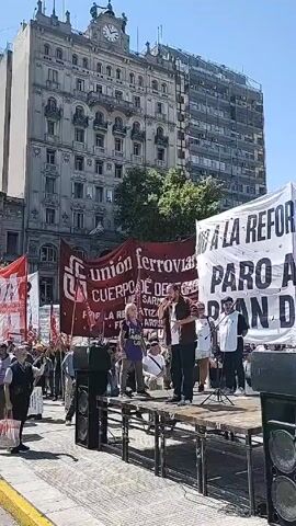 A daytime, eye-level shot depicts a political rally in an urban plaza. A crowd of people stands facing a makeshift stage where several individuals are gathered. The person at the center of the stage is speaking into a microphone, gesturing with their hands. Behind the stage are several large banners, including one reading "UNION FERROVIARIA" and another with "NO A LA REFORMA." The crowd is holding flags and banners, including one reading "IZQUIERDA SOCIALISTA" and another with "POLO OBRERO." The architecture in the background includes a tall building with a clock tower, and several other buildings with varying designs. The overall scene suggests a political demonstration or protest. The environment is sunny and bright.