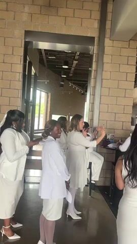 The video captures a group of approximately eight women, most of whom are dressed in white dresses or white outfits, gathered indoors. The setting appears to be a modern church or community center, evidenced by a large wooden cross and a sign reading "CROSSPOINT" in the background, with "New Here" also partially visible. A prominent brick column is in the foreground, and large windows are seen in the distance, indicating it is daytime. One woman in the center, wearing a white dress and white high heels, is holding a smartphone up, seemingly taking a picture or video. The women are interacting and posing, suggesting a social gathering or special event where white attire is worn. Another woman in white is seated further back, and several other women in white are conversing in the background. The atmosphere appears bright and communal.
