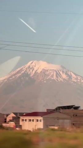 The video shows a time-lapse view of a snow-capped mountain, likely Mount Ararat, as seen from a moving vehicle. The mountain is visible in the background, with its peak covered in snow and its slopes showing patches of rock and snow. In the foreground, there are some industrial buildings and a tall transmission tower, suggesting the view is from an urban or industrial area. The sky is clear and light blue. Reflections of what appear to be power lines and the vehicle's interior are visible on the surface through which the video is filmed, likely a window. The light suggests it is either sunrise or sunset, casting a warm glow on the mountain. The camera angle is static, but the slight movement of the foreground elements implies the viewpoint is in motion. There are no people or animals visible in the frame.