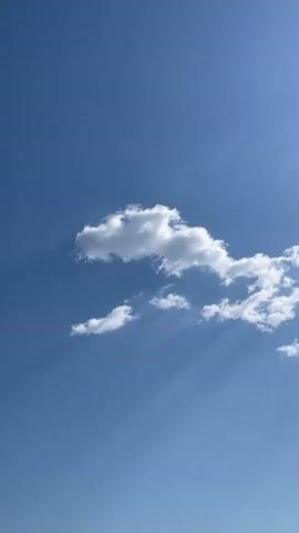 This is a video file of a clear blue sky with scattered white clouds. The sun is shining, casting rays of light through the clouds, creating dramatic light beams. There are a few wispy clouds in the foreground, and a larger, more defined cloud formation in the mid-ground. In the distance, there are faint hints of land or hills, suggesting a natural landscape. The overall impression is one of a bright and peaceful day. The video appears to be filmed during daylight hours, likely late morning or early afternoon, given the angle of the sun and the intensity of the light. There are no people or man-made objects visible in the frame, focusing solely on the natural beauty of the sky and clouds. The absence of any text or overt action contributes to a serene and contemplative mood. The location context provided indicates this scene is in Sevlievo, Bulgaria.