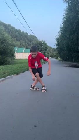 A young boy in a red shirt and black shorts is shown on an asphalt road. He is in the process of kicking a soda bottle. The bottle is on the ground and the boy is in mid-kick, his body blurred with motion. He is wearing sandals. In the background, there is a tree line, a house with a green roof, and a power line running overhead. The sky is a pale blue.