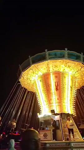 The video displays a spinning amusement park ride at night. The ride is illuminated by numerous yellow and orange lights, with decorative elements on the central structure. Several people are visible on the ride, seated in swings that are moving in a circular motion. The background is dark, indicating nighttime. People are also present at the base of the ride, some observing.