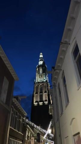 The video shows a tall, illuminated tower rising into a deep blue twilight sky. The tower is framed by buildings on either side of a narrow street. The buildings on the left appear to be brick, while the buildings on the right are painted white. The street is decorated with lights. A small crescent moon is visible in the sky. A voice can be heard in the background.