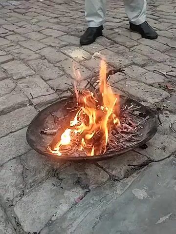 On a brick-paved surface, a small fire burns inside a shallow, round, metal bowl. The fire is composed of small sticks and twigs, producing bright orange flames. A person wearing black sandals and black pants stands behind the bowl. The overall environment appears to be outdoors, potentially a courtyard or walkway.
