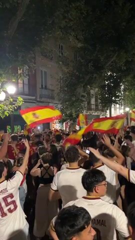 A dense crowd of people are gathered on a street at night, many holding and waving Spanish flags. Most individuals are wearing white t-shirts with red trim and what appear to be names and numbers on the back. One person in the foreground wears a t-shirt with "RAMOS 15" visible. Another person in the mid-ground wears a t-shirt with "CUENCA 24". The atmosphere appears celebratory with hands raised in the air. Streetlights and building facades are visible in the background.
