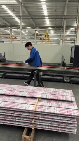 The video depicts the inside of a factory. Initially, a man in a blue uniform walks past a stack of what appear to be metal sheets, towards a machine. The camera then pans to show a group of people gathering near the metal sheets. One man is holding up a phone, possibly showing something to the group. Several people are casually dressed while others are in work uniforms. In the background, there are more workers, machines, and general factory equipment under a bright industrial ceiling.