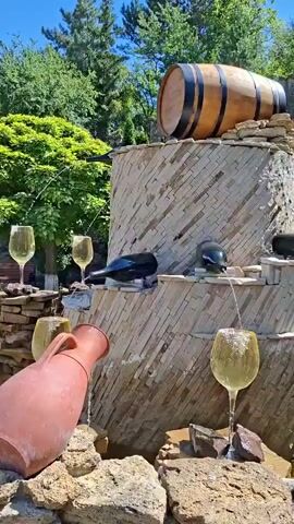A fountain designed to resemble wine is featured. A wooden barrel rests atop a tiered stone structure. Multiple dark wine bottles are strategically placed on the tiers, appearing to pour liquid into large wine glasses below. The liquid flows from the bottles, down the stone tiers, and fills the glasses. In the foreground, a terra cotta jug also appears to be part of the water feature, pouring liquid into a glass. The fountain is situated outdoors, with lush green trees and foliage visible in the background. The sky is a clear blue. The overall impression is a playful and thematic water display.