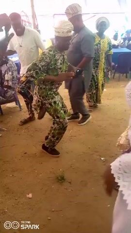 The video shows several people dancing outdoors under a tent. The ground is dirt. One person, wearing a green and brown patterned outfit and a white hat, dances actively. Other people stand around, some watching and one other dancing. The people are all wearing casual clothing. The tent has white draping. Chairs and tables are visible in the background. The video is filmed with a phone camera. The word “TECNO” and a logo appear in the lower left corner.