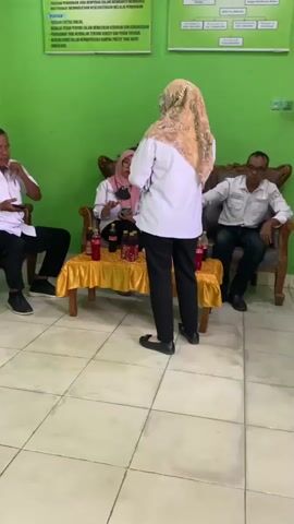 The video shows a group of people in a room with green walls and tiled floors. In the foreground, a person in a white shirt and black pants stands facing away from the camera, appearing to be talking to someone seated at a table covered with a green tablecloth. To the left of the table, a woman in a hijab and a patterned top sits with her hands clasped. Further left, a man in a gray collared shirt and black pants sits on a red chair. To the right of the table, a man in a white polo shirt with a logo and black pants sits on a chair, holding a phone. A small table with a yellow tablecloth is set between the man in the white polo and the woman at the table, with red bottles and cups on it. In the background, two framed posters are visible on the green wall.