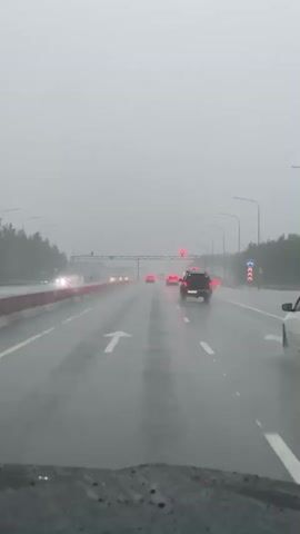 The video shows a view from inside a vehicle driving on a wet highway during heavy rain. The road is visible with lane markings and other vehicles, including cars and possibly a truck. The weather appears overcast and foggy, with reduced visibility. The driver's side windshield has raindrops on it, causing some distortion. The video captures the movement of traffic on the highway under adverse weather conditions.