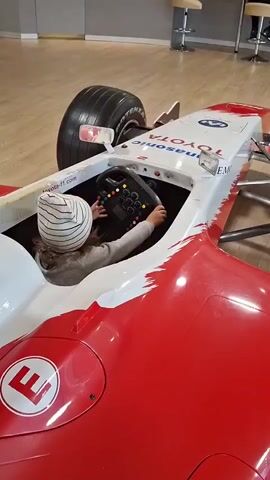 A child wearing a striped beanie is sitting in the cockpit of a red and white Formula 1 racing car replica. The child is holding the steering wheel, which has multiple buttons and controls. The car has "TOYOTA" and "Panasonic" logos visible on its body. The car is on a light-colored wooden floor, and in the background, there are bar stools and a counter, suggesting an indoor public space like an exhibition or a showroom. The lighting is bright and even.
