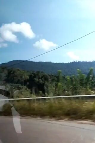 This is a video of a vehicle moving along a road. The camera is looking out from the vehicle. On the left side of the frame, there is a white building with a grey roof, surrounded by trees. In the background, there are lush green hills covered in trees. The sky is blue with some scattered white clouds. As the vehicle moves, the landscape changes slightly, showing more dense vegetation and some power lines running across the frame. The road appears to be paved, and there is a concrete barrier along the side of the road. The overall impression is of a scenic drive through a rural, tropical or subtropical area.
