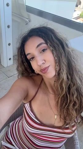 The video begins with a close-up selfie of a young woman with long, wavy brown hair and braids framing her face. She is wearing a striped dress with thin straps and a delicate necklace. She smiles and turns her head slightly, revealing a glimpse of a balcony setting behind her. The camera then pans out to show a wider view of the balcony with a railing and a potted plant. In the background, a historic fortress-like building is visible under a clear blue sky, with other buildings and a body of water in the distance. The video captures a moment of relaxation and scenic enjoyment.