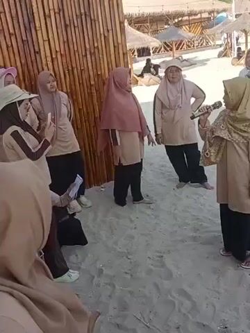 A group of women in hijabs are on a sandy beach. One woman is holding a microphone and speaking to the group. The other women are listening, some are smiling, and one is taking a photo with her phone. There is a bamboo wall behind the women, and beach umbrellas and seating areas are visible in the background. The overall atmosphere appears to be casual and friendly.