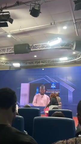 The video shows an indoor event in what appears to be a conference hall. Several rows of blue seats are occupied by attendees who are facing a stage. On stage, a large screen displays a video of a man in a pink shirt and traditional cap speaking. The event logo on the screen reads "YOUTH EMPLOYMENT: SKILLS OVER DEGREE." To the right of the screen, three people are seated, seemingly part of a panel or discussion. The ceiling is equipped with lighting fixtures and a truss structure. The camera is positioned behind the audience, offering a partial view of the attendees and focusing on the screen and stage.