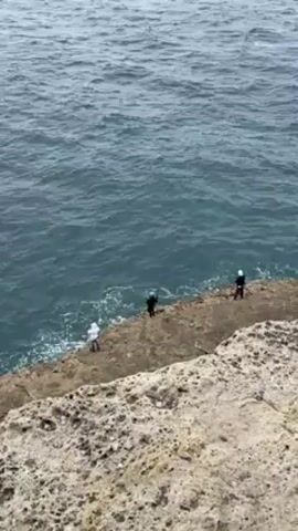 The video shows a group of four individuals standing on a rocky coastline adjacent to the ocean. The camera is positioned at an elevated angle, looking down upon them and the sea. The ocean surface is visible with small waves, and the sky is overcast. The rocky land is gray and rugged, and the individuals are spaced apart along the edge, appearing to be fishing.