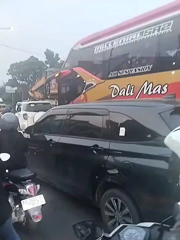 The video shows a traffic jam on a street. There are cars, trucks, and motorcycles all waiting in line. A large red and gold bus labeled "Dali Mas" is prominent. The sky is overcast. The camera is moving slightly, likely held by someone on a motorcycle. People are wearing helmets.