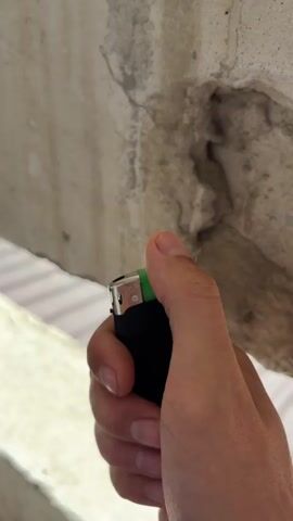 A close-up shot shows a person's hand holding a black and green lighter. The lighter is in use, with a small flame igniting from the top. The background is blurred, but appears to be an outdoor setting with a concrete structure in the upper portion of the frame. The concrete is rough and weathered, with some discoloration and a hole visible. The lower part of the background is lighter and appears to be a fence or railing. The lighting suggests it is daytime, with natural light illuminating the scene. The focus is on the hand and the lighter, with the flame being a prominent element. There is no discernible text in the image.