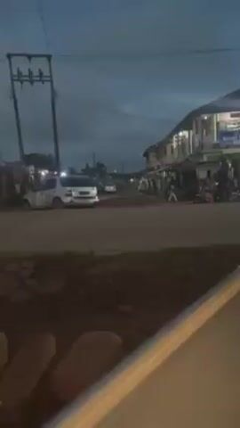 The video shows a street scene in what appears to be a small town or urban area, likely in the early morning or late evening, judging by the lighting. There is a white SUV parked on the side of a road with several other vehicles in the background. A few individuals are walking along the street, and the buildings along the roadside appear to be a mix of residential and commercial structures. Power lines are visible overhead. The road surface seems to be somewhat uneven, with patches of exposed dirt or gravel, which indicates that the road may be under construction or in need of repair. A motorcyclist passes by.
