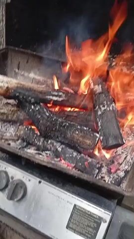 The video shows a close-up view of a fire burning in a grill. The flames are bright orange and yellow, consuming logs of wood that have turned black and are covered in ash. Smoke rises from the fire. The grill itself appears to be made of metal, with some controls visible on the front. The background is dark, suggesting the video was shot at night or in a shaded area. The overall impression is one of intense heat and the raw power of fire.