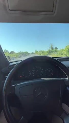 This video shows a first-person perspective from inside a Mercedes-Benz car. The driver's hands are visible on the steering wheel, and the dashboard instruments are in view. The car appears to be in motion on a paved road, with a clear blue sky and green trees and vegetation on either side of the road. The sun is shining, suggesting it is daytime. The road ahead curves gently to the right. The speedometer shows the car is traveling at approximately 80 kilometers per hour. There are no other vehicles or people clearly visible in the frame. The video appears to be a brief recording of a drive.