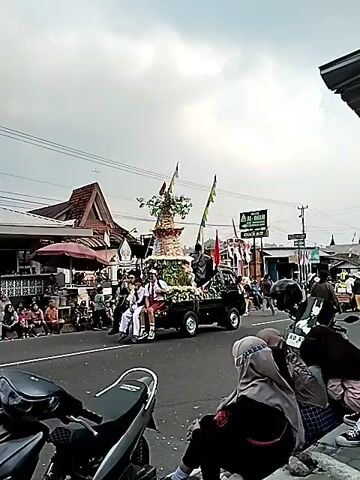 A street parade is in progress. A truck with a decorative float is moving down the road. The float is adorned with a tiered structure that resembles a small building or tower, topped with a tree. Various fruits and vegetables are incorporated into the float's decoration. Two individuals, possibly students in uniform, are seated on the front of the truck. Several other people are visible on the truck, some in uniform. On the sides of the road, a crowd of spectators is gathered, sitting and standing, observing the procession. Buildings and shops line the street. The sky is overcast. Flags and banners are displayed throughout the scene, adding to the festive atmosphere. A motorcycle is parked in the foreground.