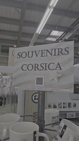 The video shows a retail display in what appears to be a store. In the foreground, there are stacked white ceramic mugs on a tiered display. Some of the mugs have text and graphics on them. A sign at the top reads "SOUVENIRS CORSICA" with a sandy background and a starfish graphic. Behind the mugs, there are shelves with glass bottles and a plush toy with a tag that says "CORSICA". The environment looks like an indoor retail space with overhead lighting and shelving units. The camera pans slightly to show different parts of the display. There are no people visible in the video.