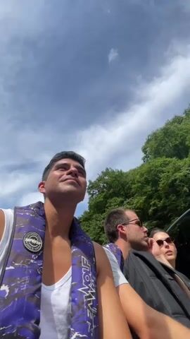 The video begins with a close-up of a young man wearing a white tank top and a purple and black life vest, looking up at the sky. The camera then pans to show other people in what appears to be a boat. There are three other individuals visible in the boat, two men and one woman. The boat is moving down a river, surrounded by lush green trees on steep hillsides. The water is a vibrant green color. The video continues to show the boat moving through the river, with a man in the back rowing with a long oar. The sky is partly cloudy. The overall scene depicts a scenic boat ride through a natural landscape.