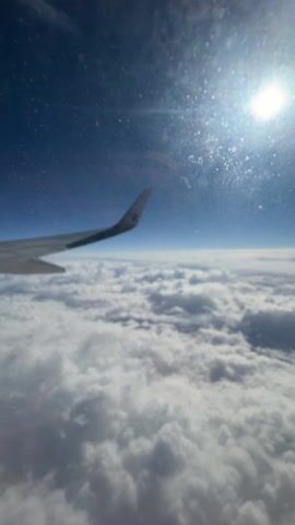 The video shows an airplane wing and clouds seen through an airplane window. The airplane wing is gray. The clouds are white and fluffy. The sky is blue. The window is dirty with spots.