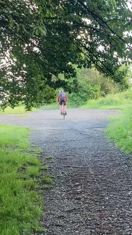 A person cycles away from the camera on a gravel path that curves to the right. The person is wearing a red cycling jersey and black cycling shorts, with a white helmet. They are riding a bicycle with a water bottle visible on the frame. The path is bordered by green grass on the left and the right. Lush green trees and foliage are visible in the background, with the sky visible through the leaves of a tree in the foreground on the left. The light suggests it is late afternoon or early evening.