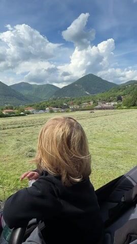 A young, fair-haired child sits in a stroller. The child is wearing a black sweater, and they are facing a field of cut grass with mountains in the background. The sky is blue with white clouds. The child turns around, smiles, takes a drink from a plastic water bottle, and then turns to face the view again.