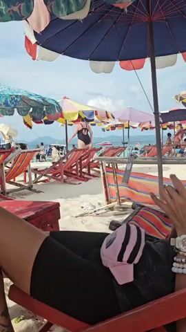 A person is lying on a red beach chair under a large umbrella, holding a mobile phone. In the background, a crowded beach with many umbrellas and lounge chairs is visible. The sea and distant islands are also in view, under a partly cloudy sky.