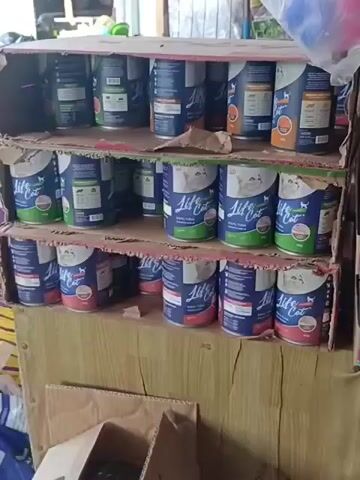 The video shows a close-up, eye-level view of shelves stocked with pet food. The shelves are made of cardboard and appear to be in a retail setting, possibly a pet store or a small market stall.

On the shelves, there are multiple rows of canned pet food, primarily cat food. The cans are predominantly blue and white, with some featuring images of cats. Brands visible include "Let's Eat" and "Whiskas." Some cans are stacked in rows, while others are arranged more casually. There are also pouches of wet cat food in pink packaging with images of cats.

In the background, further shelves are visible, stocked with various dry goods in bags, possibly grains or other pet food ingredients. The overall environment appears somewhat cluttered but organized for display. The lighting suggests an indoor setting, possibly with natural light from an unseen source. No people are visible in the frame.
