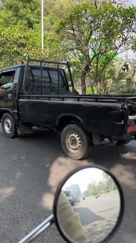 A black Mitsubishi pickup truck drives on a paved road. In the foreground, a motorcycle's side mirror reflects a street with other vehicles. The truck's bed has a white logo that reads "MITSUBISHI." Further down the road, two people on a white scooter pass by. To the right of the road, several trees with green foliage are visible, and behind them, a building with a green roof can be seen. The environment appears to be an urban or suburban setting with a clear sky.