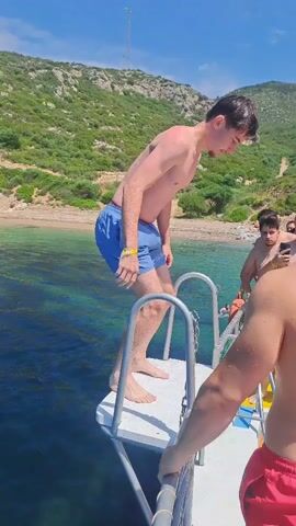 A young man, wearing blue swim trunks, stands on a white platform at the back of a boat, preparing to jump into the clear, blue-green water. He has short dark hair and a light complexion, with his bare chest visible. The platform has metal railings. In the background, a lush green hillside rises from a sandy and rocky coastline under a bright blue sky, indicating a sunny day.

As the video progresses, the man leans forward, then executes a clean backflip, rotating completely before entering the water. A large splash is created where he enters the water, with white foam and spray momentarily obscuring the view of him. The surrounding water shows varying depths with darker patches indicating underwater features, possibly seaweed or rocks. Another person in red swim shorts is partially visible on the right side of the frame, near the boat. The scene suggests a recreational activity, likely a boat trip or swimming excursion in a coastal area, specifically Menderes, Turkey, as provided in the context.