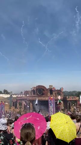 The video captures a festival scene with a large, elaborate stage structure in the background. The stage appears to be designed to resemble ancient ruins, with detailed stonework and ornate decorations. A crowd of people is gathered in front of the stage, many holding colorful umbrellas, including a prominent pink and a yellow one in the foreground. The sky is a clear blue with wispy white clouds and what appear to be trails from fireworks or pyrotechnics. Confetti or streamers are visible falling from the stage, and there are bursts of colored smoke or light emanating from the stage as well. The overall atmosphere suggests a lively outdoor event, possibly a music festival, with a performance or show in progress.