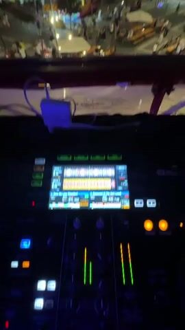 This video appears to be taken from a raised platform, possibly a DJ booth, overlooking a lively outdoor event at dusk. In the foreground, DJ equipment is visible, including a console with a screen displaying waveforms and controls. The background reveals a bustling town square filled with people, tents, tables, and lights. An ornate building, likely a church, stands prominently on the left side of the square. Numerous people are scattered throughout the area, some standing, others seated at tables. Several string lights are strung across the square, illuminating the scene. The atmosphere suggests a festive gathering, perhaps a market or a local festival. The camera pans slightly, capturing different sections of the crowded square and the darkening night sky above.