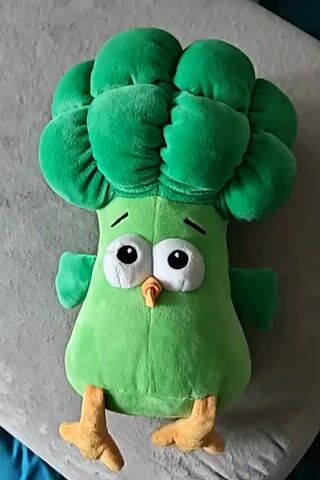 A plush toy shaped like a green broccoli with large eyes and orange feet is lying on a gray surface. The toy is positioned vertically with its head at the top. The background consists of a blurred gray surface and hints of a blue fabric.