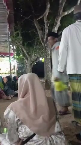 The video shows a group of people gathered outdoors at night, possibly for a religious ceremony or event. In the foreground, several individuals are seated, facing away from the camera, some wearing hijab. To the right, a man in a white shirt and sarong stands facing away. In the mid-ground, a man in a light-colored jacket and cap walks from right to left, carrying a yellow bag. Further back, a stage is visible with people seated on it, illuminated by lights. The environment appears to be a rural or semi-outdoor setting with trees and a makeshift canopy overhead. The lighting is dim, with artificial lights illuminating the stage and surrounding areas.
